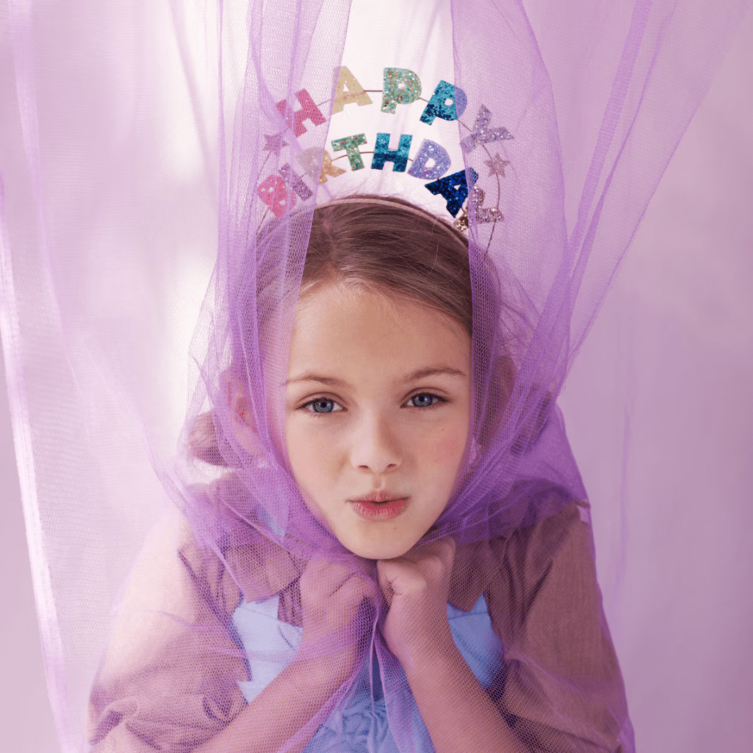 Multicolor happy birthday tiara with glitter letters worn by child under purple tulle for birthday from Parte Haus party supply store in Bethesda Maryland